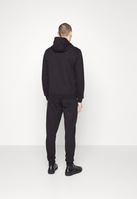 Svart hoodie och matchande byxor i ett mjukt, texturerat tyg, med huva och ribbade ärmslut, tillsammans med svarta skor.