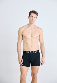 Svarte boxertruser med en hvit "HILFIGER" logo på midjebåndet, laget av mykt materiale, med en tettsittende passform og design som går til midten av låret.