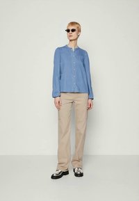 Personne portant une blouse bleu à boutons, un pantalon cargo beige, des chaussures argentées épaisses et des lunettes de soleil noires en forme d'œil de chat sur un fond clair.