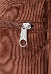 Καφέ κοτλέ ύφασμα με κατακόρυφες ραβδώσεις, που διαθέτει φερμουάρ σε ασημί χρώμα με το λογότυπο "JS". Η κοντινή λήψη υπογραμμίζει την υφή και τις λεπτομέρειες του σχεδιασμού.