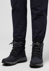 Scarponi da trekking neri con lacci grigi, colletto imbottito, pannello laterale testurizzato e suola robusta, abbinati a pantaloni blu navy.