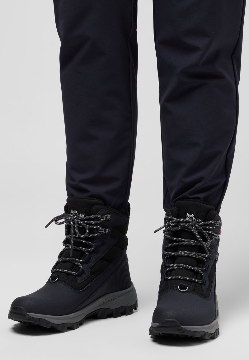 Scarponi da trekking neri con lacci grigi, colletto imbottito, pannello laterale testurizzato e suola robusta, abbinati a pantaloni blu navy.