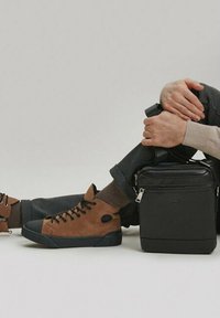 Bruine suède high-top sneakers met zwarte rubberzolen, gecombineerd met donkere broeken. Een stijlvolle zwarte vierkante tas staat naast de benen van de drager.