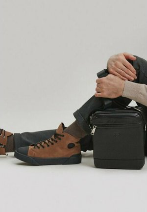 Brązowe zamszowe sneakersy typu high-top z czarnymi gumowymi podeszwami, w połączeniu z ciemnymi spodniami. Elegancka czarna kwadratowa torba leży obok nóg użytkownika.