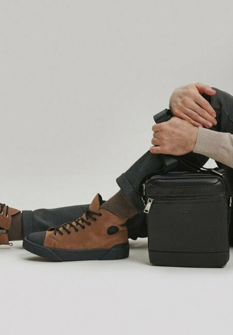 Bruine suède high-top sneakers met zwarte rubberzolen, gecombineerd met donkere broeken. Een stijlvolle zwarte vierkante tas staat naast de benen van de drager.