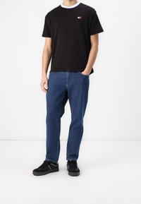 Nuori mies, jolla on päällään musta t-paita, siniset farkut ja mustat tennarit, seisoo valkoista taustaa vasten, toinen käsi taskussa.