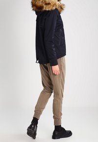 Tummansininen takki, jossa on tekoturkishuppu, vetoketjullinen etutasku ja joustavat hihansuut, yhdistettynä vaaleanbeigeihin kapeneviin housuihin ja mustiin jalkineisiin.