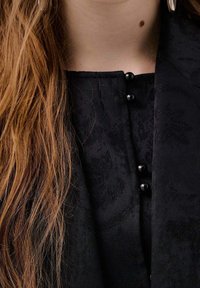 Musta mekko tekstuurilla, jossa on pyöreä kaula-aukko ja viisi mustaa nappia edessä. Pitkä ruskea hius osittain näkyvissä.