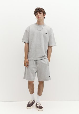 Junger Mann trägt ein hellgraues oversized T-Shirt und passende Shorts, weiße Socken, braune Sneakers, steht vor einem einfarbigen weißen Hintergrund.