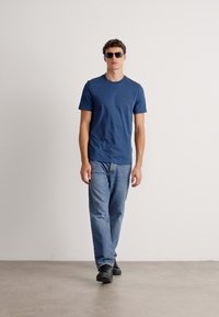 Hombre caminando hacia adelante, vistiendo gafas de sol oscuras, camiseta azul, pantalones vaqueros claros y zapatos negros, contra un fondo claro y simple.