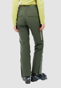 Groene waterdichte skibroek met een getailleerde pasvorm, achterzakken en ritsventilatie aan de zijkanten; gedragen met zwarte skikleding met blauwe accenten.
