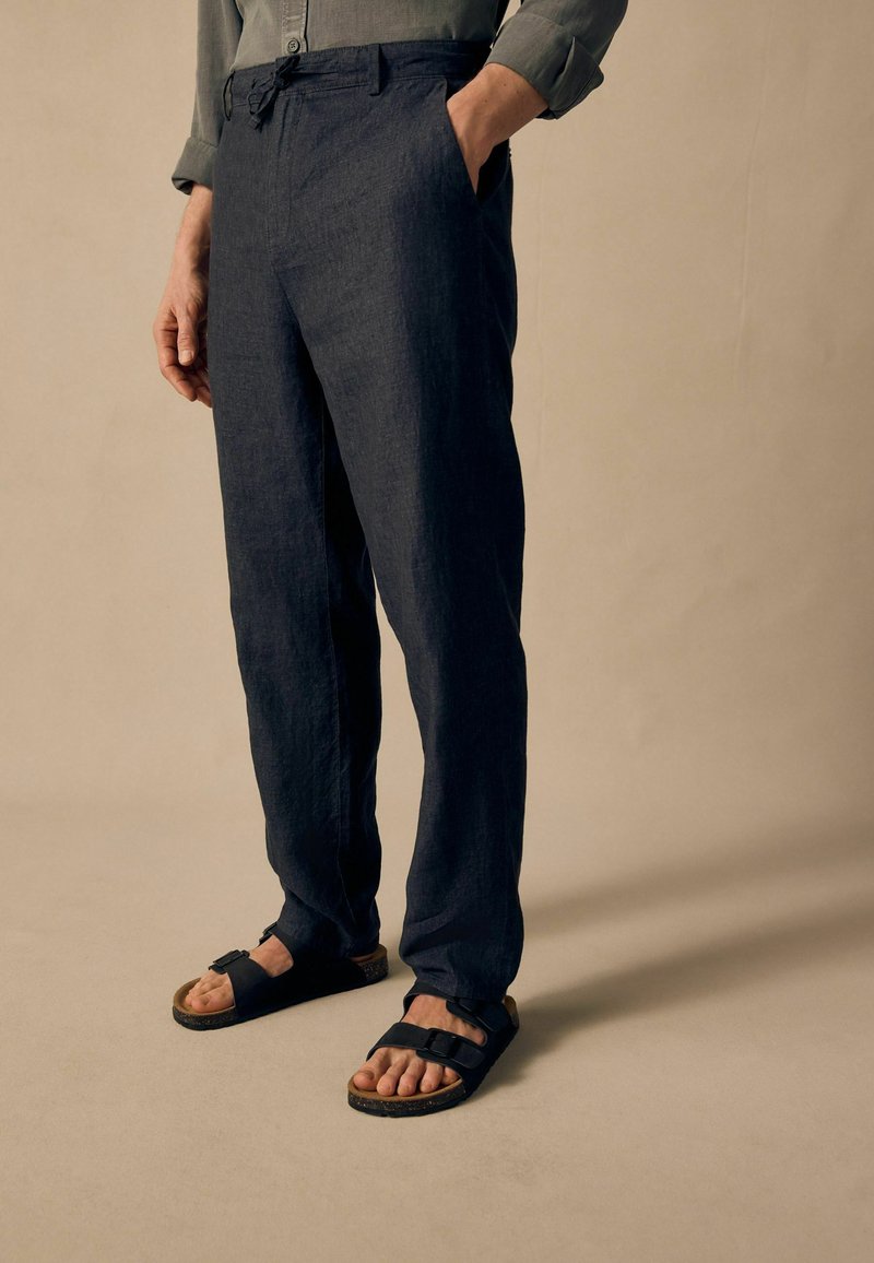 Pantalons en lin léger bleu marine foncé avec une coupe décontractée et des passants de ceinture. Associés à des sandales noires à deux brides sur pieds nus.