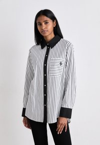 Svart og hvit stripete skjorte med svart krage og knappestolpe, har brystlomme og en liten logo. Stoffet ser glatt ut.