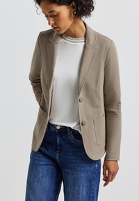 Ljusbeige blazer med skräddarsydd passform, som har två framfickor, slagkrage och en mjukt strukturerad silhuett, bärs över en vit topp.