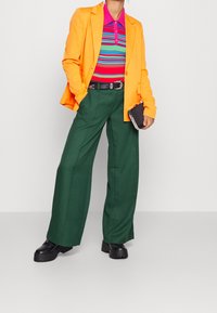 Orange kavaj, randig polo i rosa, grön och blå, gröna vida byxor, svarta kraftiga skor, håller i en liten svart kuvertväska.