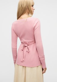 Γυναίκα με μακριά ξανθά μαλλιά, ντυμένη με ροζ μακρυμάνικη μπλούζα δεμένη στην πλάτη και μπεζ φούστα, στραμμένη προς τα πίσω σε απλό φόντο.