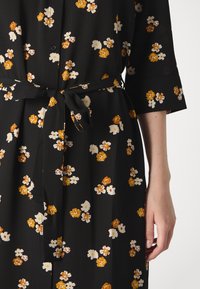 Mustrine lilledega must kleit, millel on vöö, kollaste ja valgete lillemuster. Materjal on tekstureeritud ja sellel on kolmeveerand-sleeved varrukad.