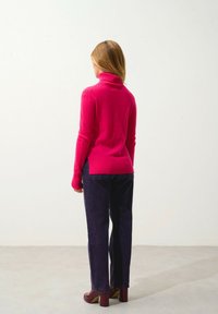 Fuksia neulepusero, jossa on korkea kaulus, yhdistettynä tummansinisille farkuille ja burgundin värisiin korokepolkkaisiin, kuvattuna takaa vaaleaa taustaa vasten.