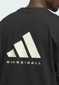 Sort t-shirt med et stort hvidt Adidas-logo og ordet "BASKETBALL" i grå under, lavet af blødt bomuldsstof.