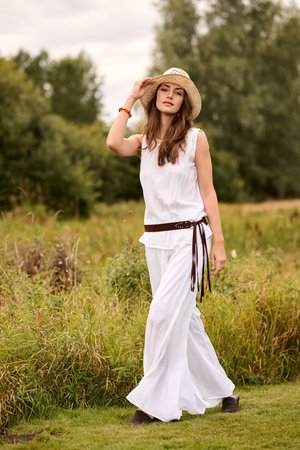 Donna con top bianco senza maniche e pantaloni a gamba larga, che indossa un cappello di paglia e una cintura marrone, in piedi in un ambiente esterno erboso con alberi.
