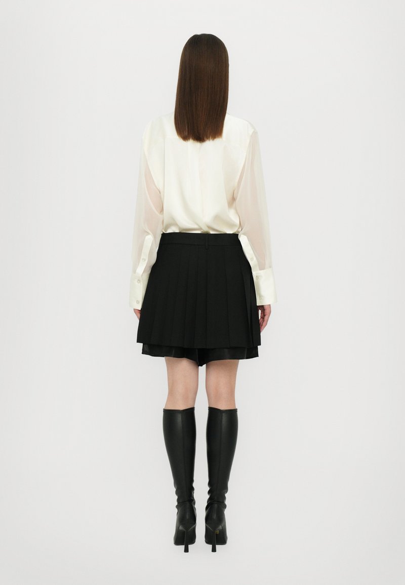 Blouse blanche avec des manches translucides, associée à une jupe plissée noire et des bottes à talons montantes noires. Design simple et élégant.