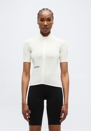 Kobieta w dopasowanej białej koszulce kolarskiej z krótkim rękawem i czarnych szortach kolarskich, stojąca zwrócona przodem na jednolitym tle.