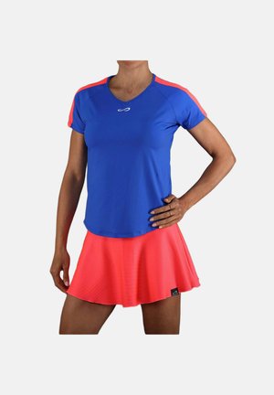 Vrouw draagt een blauw atletisch shirt met korte mouwen en rode strepen en een felrode uitlopende sportrok tegen een witte achtergrond.