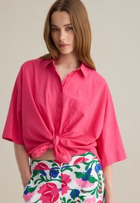 Camicia a maniche corte di un vivace colore rosa con bottoni e nodo frontale, accompagnata da pantaloni corti floreali a motivi verdi e bianchi.