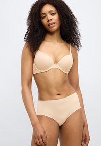 Γυναίκα με σγουρά μαλλιά που φοράει μπεζ σουτιέν και αόρατο εσώρουχο, στέκεται μπροστά σε απλό λευκό φόντο.