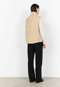Vest bej matlasat cu guler înalt, suprafață texturată, purtat peste un top deschis la culoare, asortat cu pantaloni negri și pantofi negri. Viziune din spate.