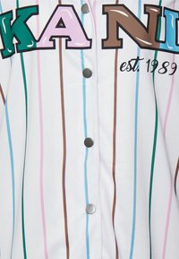 Bijela košulja s dugim rukavima s šarenim vertikalnim prugama u zelenoj, ružičastoj, smeđoj i plavoj boji; sadrži veliki crni natpis i detalj godine "osn. 1989."