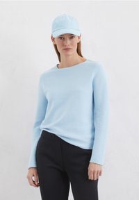 Helles blauer langärmeliger Pullover mit rundem Ausschnitt, kombiniert mit schwarzen Hosen. Eine passende hellblaue Mütze rundet das Outfit ab.