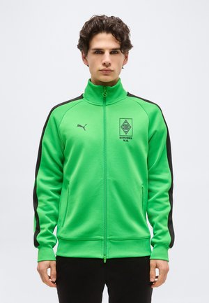 Νεαρός άνδρας που φοράει μπλούζα φερμουάρ Puma της Borussia M.G. σε έντονο πράσινο χρώμα με μαύρες ρίγες στα μανίκια, στέκεται μπροστά σε ένα απλό φόντο.