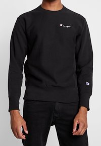 Felpa Champion nera indossata da una persona, con piccolo logo sul petto e sulla manica, abbinata a pantaloni neri su sfondo semplice.