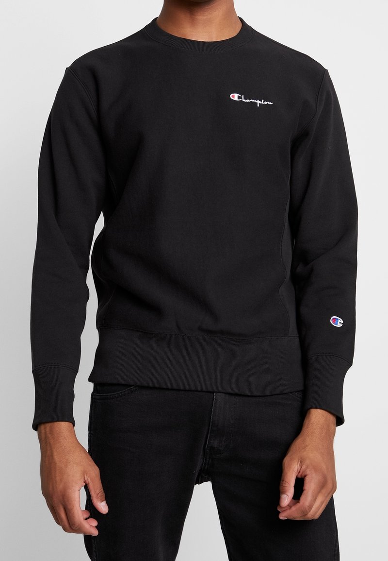 Felpa Champion nera indossata da una persona, con piccolo logo sul petto e sulla manica, abbinata a pantaloni neri su sfondo semplice.