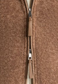 Makro záber na hnedú textúrovanú fleecovú tkaninu so stredným obojsmerným zipsom s koženými ťahadlami.