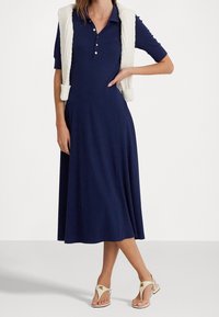 Mörkblå poloklänning med utsvängd kjol, korta ärmar och treknappsknäppning. Matchad med en vit stickad väst och beige sandaler.
