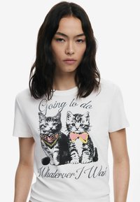 T-shirt en coton blanc avec deux chats illustrés. L'un porte un collier, l'autre un nœud papillon rose. Le texte indique "Je vais faire ce que je veux."
