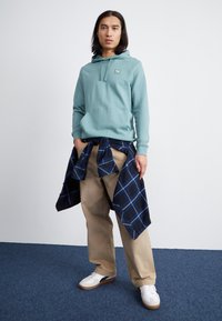 Hoodie verde menta con tasca frontale, abbinato a pantaloni beige. Camicia a quadri navy legata in vita. Sneakers bianche con suola marrone.