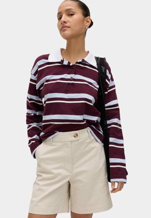 Kvinde iført en langærmet poloshirt med vinrøde og lyseblå striber, beige shorts med høj talje og en sort skuldertaske i net.