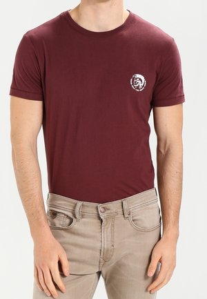 Bordowa bawełniana koszulka z krótkim rękawem i okrągłym dekoltem. Na lewej stronie klatki piersiowej znajduje się okrągłe białe logo graficzne. Noszona z jasnobeżowymi dżinsami.