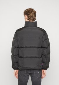 Giubbotto imbottito nero con colletto alto, caratterizzato da un design trapuntato e polsini elasticizzati. Indossato sopra jeans scuri, con un logo applicato.