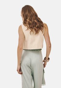 Chaleco corto beige con un diseño sencillo, combinado con pantalones de satén verde claro que tienen un borde decorativo de pompones en la cintura.