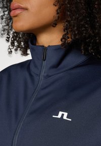 Marinblå zip-up jacka med hög krage, slät textur och en liten vit logotyp på vänster sida av bröstet.