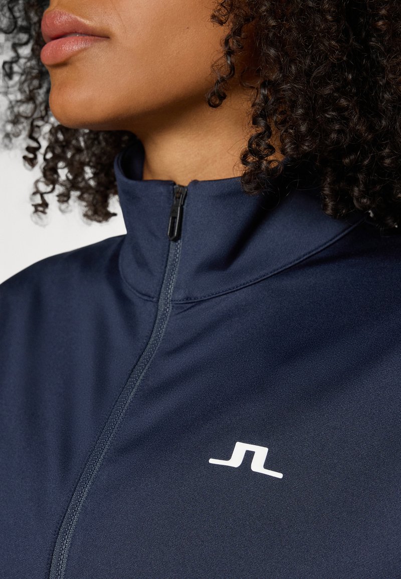 Navy zip-up jacket featuring a high collar, smooth texture, and a small white logo on the left side of the chest.