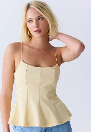 Młoda kobieta o blond krótkiej fryzurze bob, ubrana w jasnopomarańczowy top na ramiączkach i niebieskie jeansy, pozująca z jedną ręką za karkiem.
