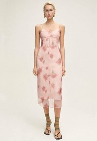 Rochie midi roz, fără măneci, cu modele florale; overlay transparent, croi drept, cu butoane subtile în față. Purtați cu sandale verzi cu barete.