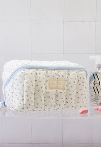 Kottont kosmetisk veske med quiltet tekstur, hvit bunn, grønt blomsterdesign, blå glidelås og en logomerke. Inkluderer hårprodukt og piller.