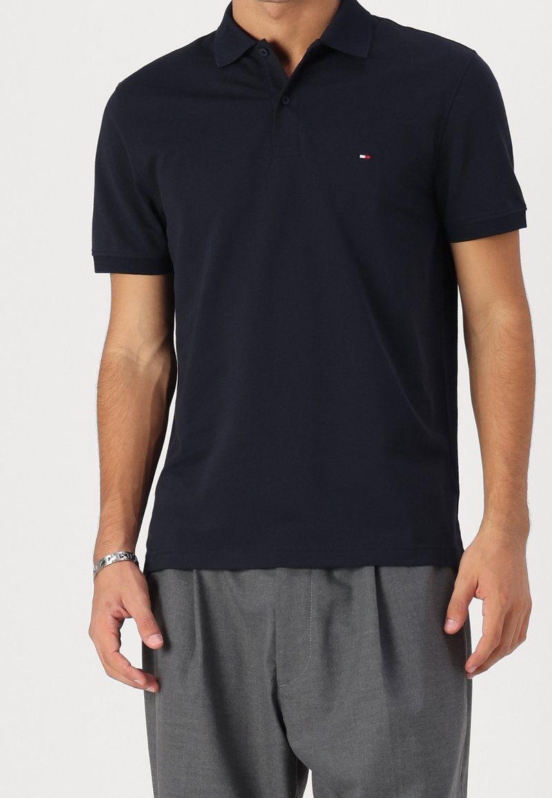 Homme portant un polo noir à manches courtes avec un petit logo sur la poitrine et un pantalon gris, debout, bras détendus.