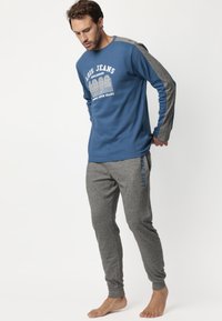 Camisa de manga larga azul con gráfico blanco y mangas grises, combinada con pantalones de chándal grises que tienen texto azul. Tejido suave, corte casual.